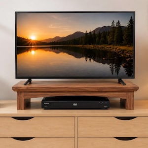 May include: A rectangular wooden monitor stand with a smooth, natural brown finish and four sturdy legs. The stand features a wide, flat top surface and an open space underneath, currently housing a black Sony DVD player on a light wood dresser.