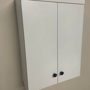 May include: White wall-mounted cabinet with two doors and black knobs.