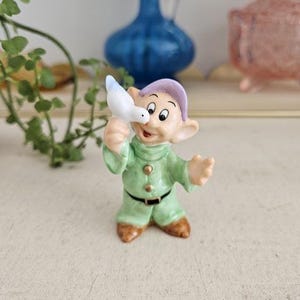 May include: A ceramic figurine of a dwarf from Snow White, wearing a green outfit with a black belt and brown shoes. The dwarf is holding a white bird. The figurine has a purple hat and is smiling.
