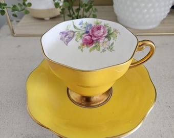 Può includere: Un set di tazza e piattino giallo con bordi dorati. La tazza presenta un motivo floreale con rose rosa e tulipani viola. Il piattino ha un bordo smerlato.