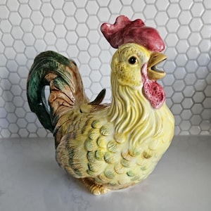 May include: A ceramic rooster ornament with a yellow body, red comb and wattle, and a green and brown tail. The rooster is depicted standing with its beak open. The ornament has a glossy finish and is likely for decorative purposes.