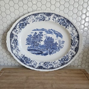 May include: Oval, white ceramic platter featuring a blue landscape design and floral border. The platter has a scalloped edge and handles. A classic serving piece.