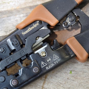 May include: Black and brown wire crimping tool with the brand name "Southwire" printed on the side. The tool has a lever and a variety of crimping slots. The tool is labeled "RJCR-T1" and has safety warnings.