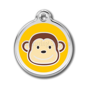 May include: A round silver pet tag with a yellow circle and a cartoon monkey face in the center. The monkey has brown fur, a white face, and a big smile.