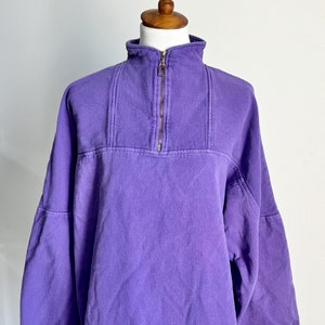 Puede incluir: Una sudadera de color morado con cremallera de un cuarto de vuelta, cuello de canalé y cremallera dorada. La sudadera tiene un corte holgado y mangas largas.