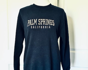 Puede incluir: Una sudadera azul marino con el texto "PALM SPRINGS CALIFORNIA" en blanco, con una fuente de estilo vintage. La sudadera se muestra en un maniquí sobre un fondo blanco.