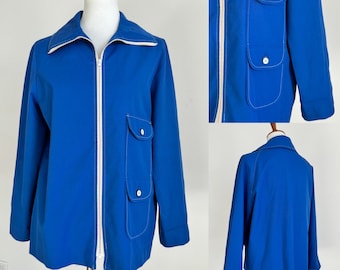 May include: A blue jacket with white piping and two pockets on the front. The jacket has a zipper closure and a stand-up collar.