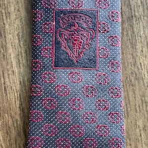May include: A patterned necktie with a repeating circular design in red and white dots on a navy blue background. The tie features a navy blue rectangular patch with a red crest and the word "GUCCI".