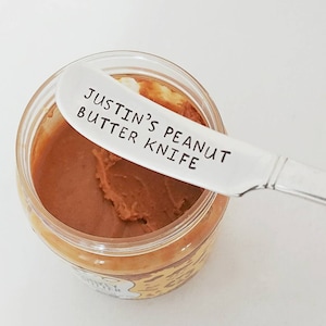 Könnte beinhalten: Ein silbernes Erdnussbuttermesser mit dem eingravierten Text "Justin's Peanut Butter Knife". Das Messer liegt auf dem Rand eines Glases Erdnussbutter.