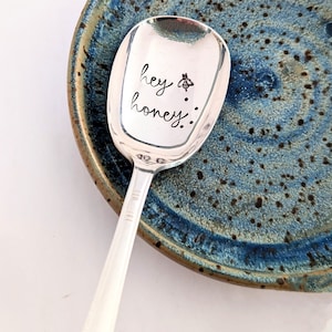 May include: A silver spoon with a blue and brown ceramic dish. The spoon has the words "hey honey" engraved on it with a small bee symbol.