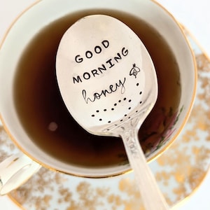 May include: A silver spoon with a decorative design and the words "Good Morning Honey" engraved on it. The spoon is resting in a teacup with a floral pattern.