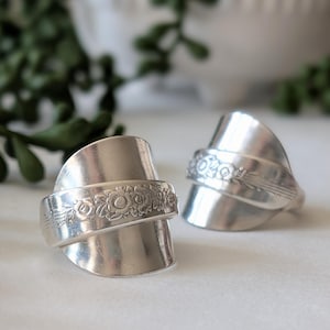 May include: Two handcrafted silver rings, each made from repurposed silverware. The rings feature a floral design on the band and a curved, spoon-like shape. The rings are displayed on a white surface.