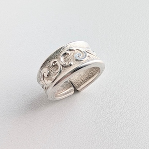May include: A silver adjustable ring with an ornate, swirling pattern. The ring features a textured surface and a small, light blue accent. The band is open and adjustable, allowing for a comfortable fit. The ring is designed to be worn on a finger.