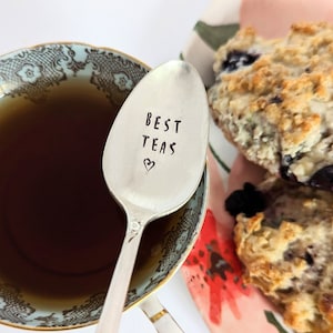 May include: A silver teaspoon engraved with the words "Best Teas" and a heart symbol. The spoon is resting in a blue and white teacup containing tea. A plate with a floral pattern and two blueberry scones is in the background.
