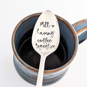 May include: A silver spoon with the words "still having coffee together" engraved on it, resting in a blue and brown coffee mug.