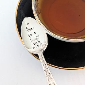 May include: A silver teaspoon with the inscription "tea is a hug in a mug" on a black and gold saucer with a teacup filled with brown tea.