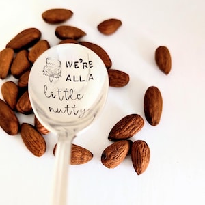 May include: A silver-toned metal spoon featuring a stamped illustration of a nutcracker and the black text "WE'RE ALL A little nutty" centred in the bowl, surrounded by a scattering of brown almonds on a plain white surface.