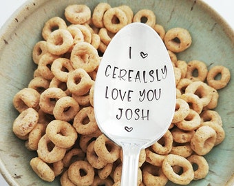 Op de afbeelding: Een zilveren lepel met de tekst "I ♥ Cerealsly Love You Josh" erop gegraveerd. De lepel ligt in een kom met ontbijtgranen.