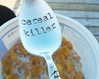 May include: A silver spoon with the words "cereal killer" engraved on it. The spoon is sitting in a bowl of cereal with milk.