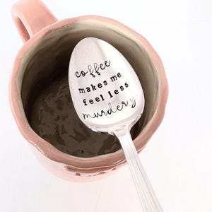 May include: A silver spoon with the text "coffee makes me feel less murdery" rests inside a pink ceramic mug. The mug is filled with a dark brown liquid. The spoon is angled towards the viewer.