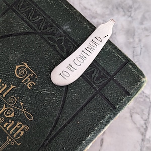 May include: A silver bookmark with the text "TO BE CONTINUED..." on a green book with gold lettering.