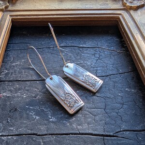 May include: A pair of silver earrings with a rectangular design and floral detailing. The earrings have long, thin hooks. The background is a dark, textured surface within a gold-colored frame.
