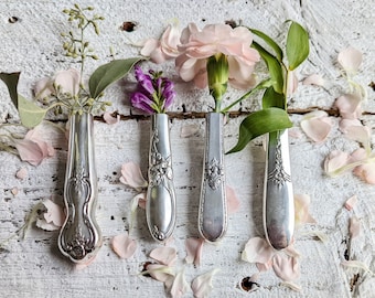 May include: Four antique silver spoon vases, each holding a different flower and greenery. The spoons are arranged on a distressed white wooden surface, surrounded by scattered pink flower petals. A unique floral arrangement.