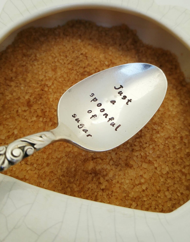 May include: A silver spoon with the inscription "Just a spoonful of sugar" resting on a bowl of brown sugar.