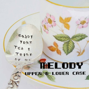 May include: A silver spoon with the inscription "Enjoy Your Tea & Think of Me" is placed next to a teacup and saucer. The teacup and saucer are white with a floral design featuring pink flowers and green leaves. The word "MELODY" is below the spoon.