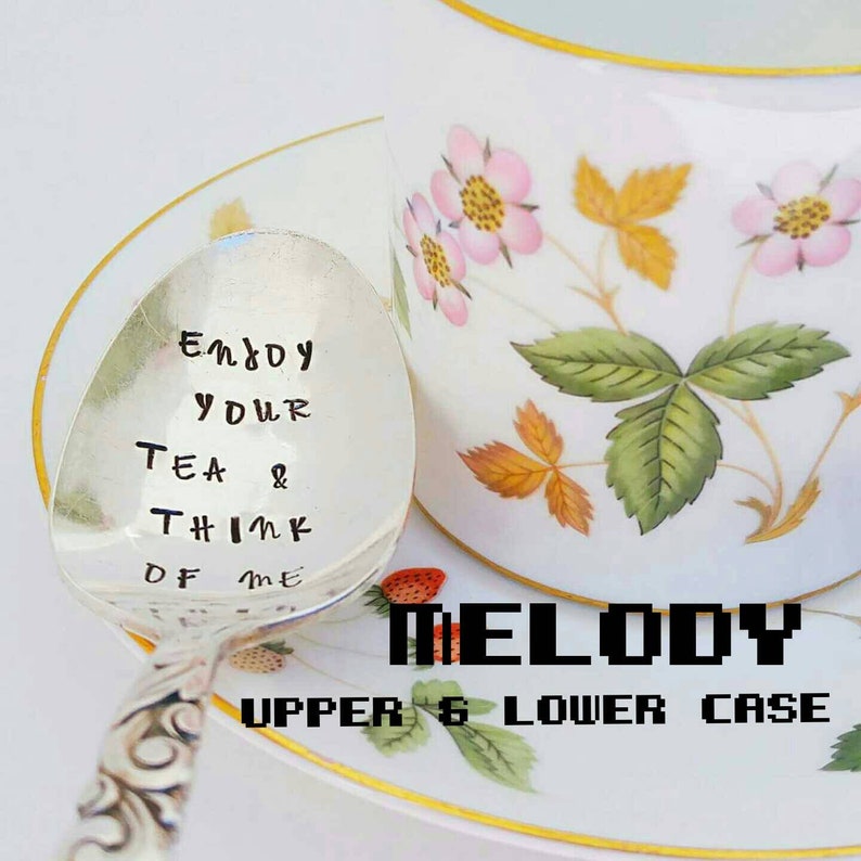 May include: A silver spoon with the inscription "Enjoy Your Tea & Think of Me" is placed next to a teacup and saucer. The teacup and saucer are white with a floral design featuring pink flowers and green leaves. The word "MELODY" is below the spoon.