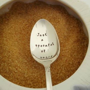 May include: A silver teaspoon with the words "Just a spoonful of sugar" engraved on the bowl. The spoon is resting in a bowl of brown sugar.