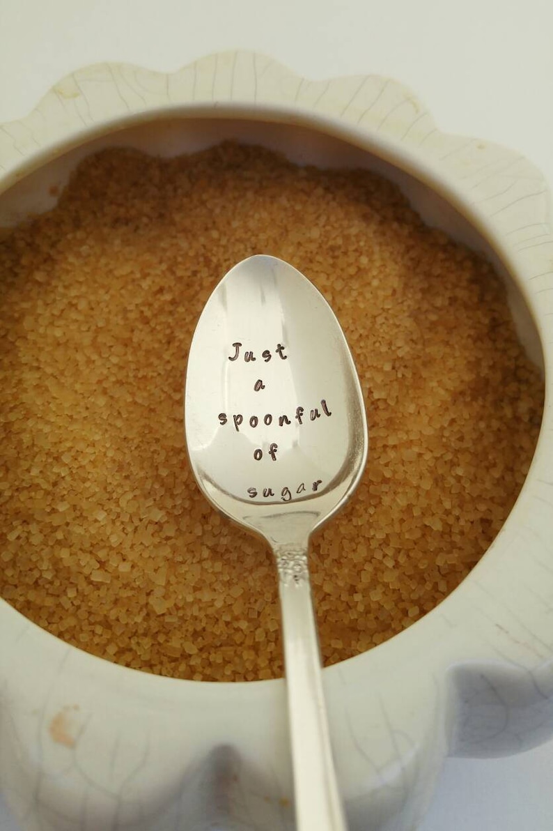 May include: A silver teaspoon with the words "Just a spoonful of sugar" engraved on the bowl. The spoon is resting in a bowl of brown sugar.
