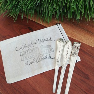 Puede incluir: Tres cucharas de plata con etiquetas grabadas: "Tomillo", "Perejil" y "Albahaca". Las cucharas est&aacute;n en una bolsa de tela blanca con las palabras "Jard&iacute;n" y "Delicias" escritas en ella.