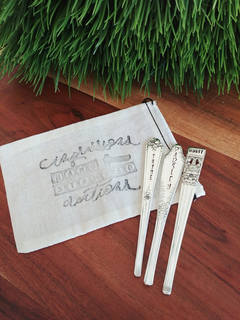 Puede incluir: Tres cucharas de plata con etiquetas grabadas: "Tomillo", "Perejil" y "Albahaca". Las cucharas est&aacute;n en una bolsa de tela blanca con las palabras "Jard&iacute;n" y "Delicias" escritas en ella.