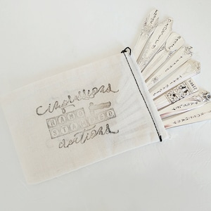 Puede incluir: Una bolsa de tela blanca con las palabras "Cimballigano Hand Stamped Creations" impresas en ella. La bolsa contiene un juego de cucharas de plata con diferentes palabras grabadas en ellas.