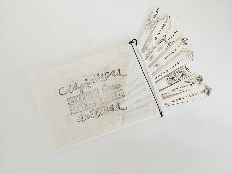 Puede incluir: Una bolsa de tela blanca con las palabras "Cimballigano Hand Stamped Creations" impresas en ella. La bolsa contiene un juego de cucharas de plata con diferentes palabras grabadas en ellas.