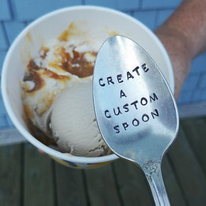 May include: A silver spoon with the words "Create a Custom Spoon" engraved on it. The spoon is resting on top of a cup of ice cream with caramel sauce.