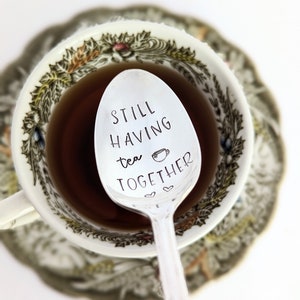 May include: A silver teaspoon engraved with the words "Still having tea together". The spoon is resting in a teacup filled with tea.
