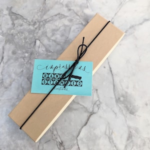 May include: A rectangular, light brown box tied with black string, with a teal tag that reads "Impressions HAND STAMPED creations" in black lettering. The box is on a marble surface.