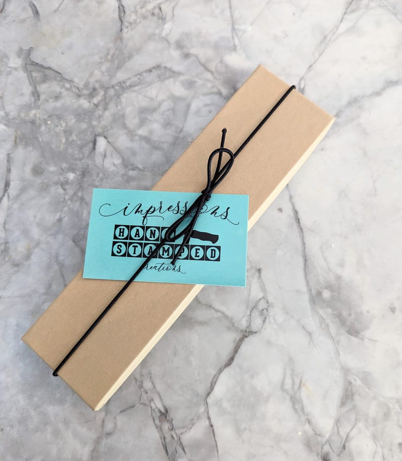 May include: A rectangular, light brown box tied with black string, with a teal tag that reads "Impressions HAND STAMPED creations" in black lettering. The box is on a marble surface.