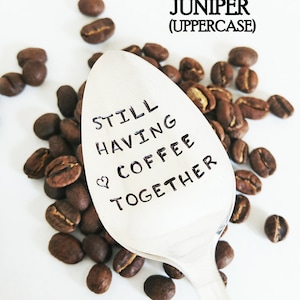 May include: A silver spoon with the text "STILL HAVING COFFEE TOGETHER" and a heart symbol, surrounded by roasted coffee beans. The word "JUNIPER" is above the spoon in uppercase letters.