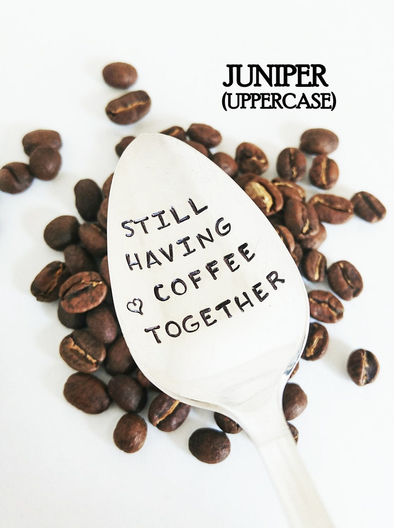 May include: A silver spoon with the text "STILL HAVING COFFEE TOGETHER" and a heart symbol, surrounded by roasted coffee beans. The word "JUNIPER" is above the spoon in uppercase letters.
