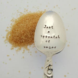 May include: A silver teaspoon with an engraved message that reads "Just a spoonful of sugar". A pile of brown sugar is next to the spoon.