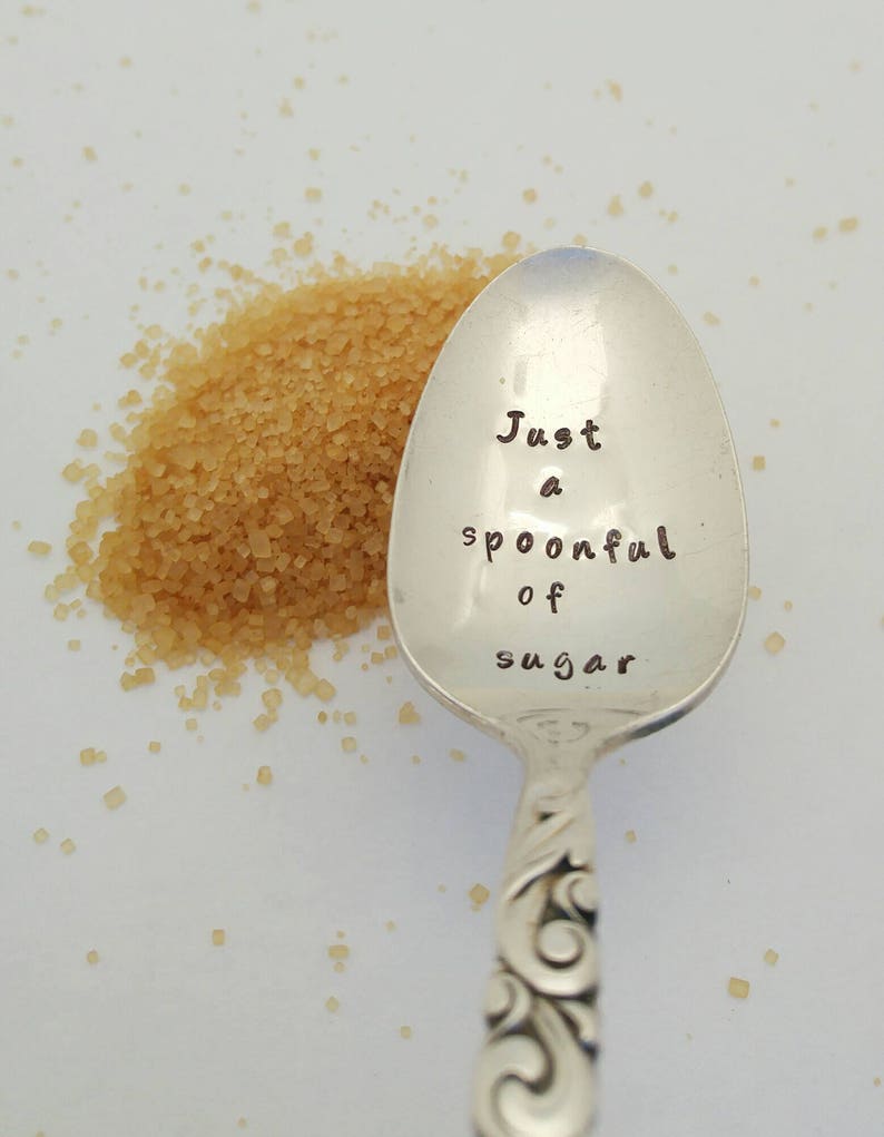 May include: A silver teaspoon with an engraved message that reads "Just a spoonful of sugar". A pile of brown sugar is next to the spoon.