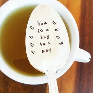 May include: A silver spoon with the phrase "Tea is a hug in a mug" and heart designs stamped on the spoon's surface. The spoon is in a white mug filled with tea, set against a wooden background.