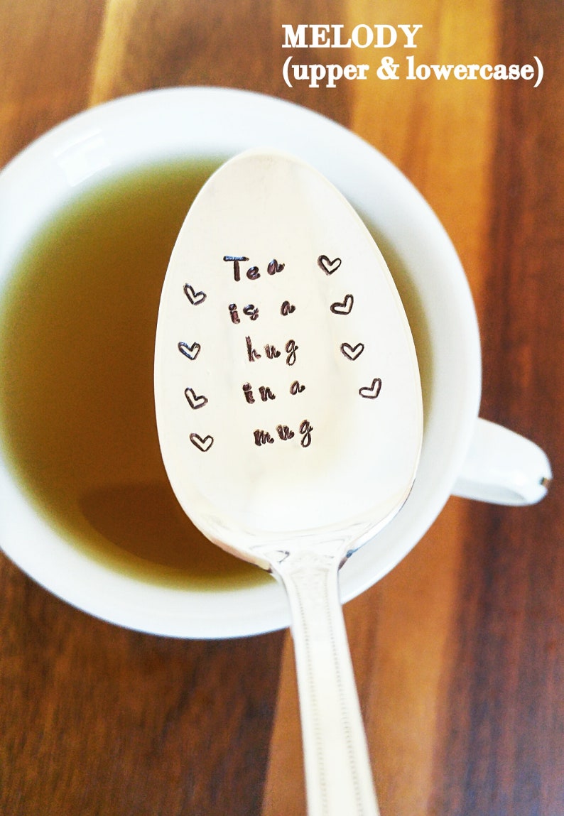 May include: A silver spoon with the phrase "Tea is a hug in a mug" and heart designs stamped on the spoon's surface. The spoon is in a white mug filled with tea, set against a wooden background.