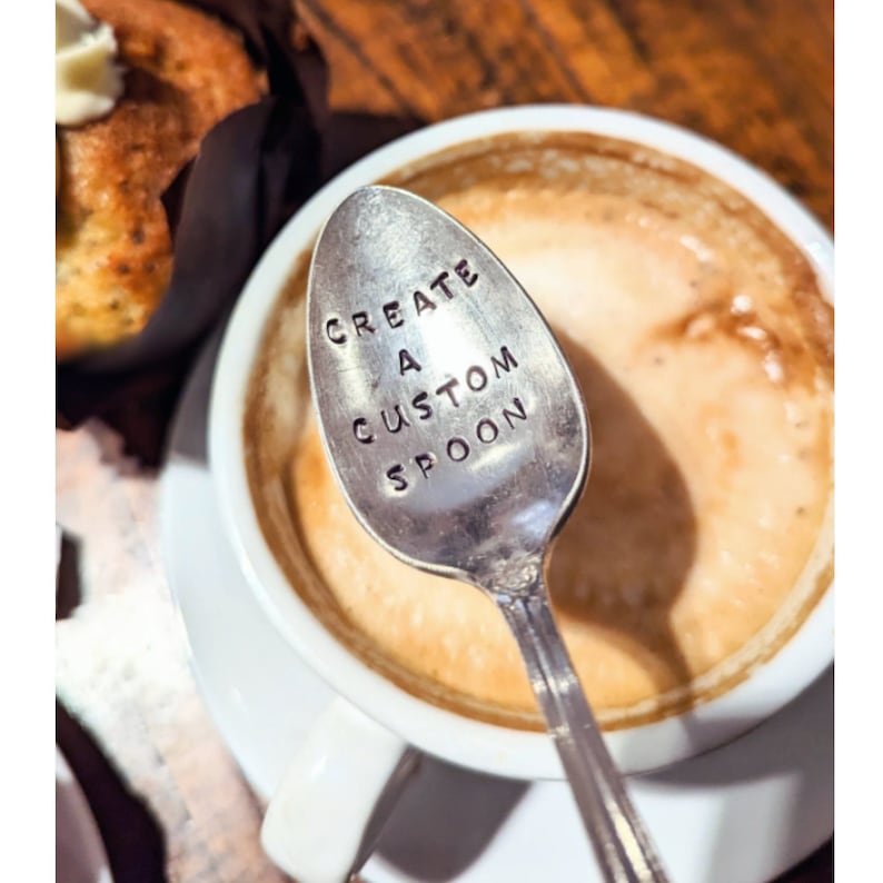 May include: A silver spoon with the words "CREATE A CUSTOM SPOON" stamped on the bowl, resting in a cup of coffee. The spoon is positioned over the coffee, with a muffin in the background.