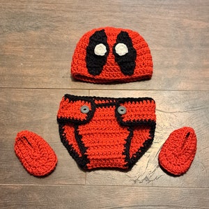 May include: A red and black crocheted baby outfit with a hat, diaper cover, and booties. The hat features a white eye mask design.