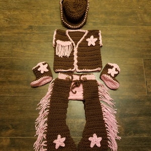 May include: A brown and pink crochet cowboy outfit for a baby, including a hat, vest, chaps, and booties. The vest has pink fringe and star accents. The chaps have pink fringe and star accents. The hat has a white trim.