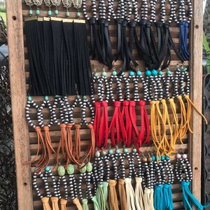 May include: A display of silver and turquoise earrings with leather fringe. The earrings are arranged on a wooden board with a slatted design. The fringe is in a variety of colors, including black, red, yellow, brown, and blue.